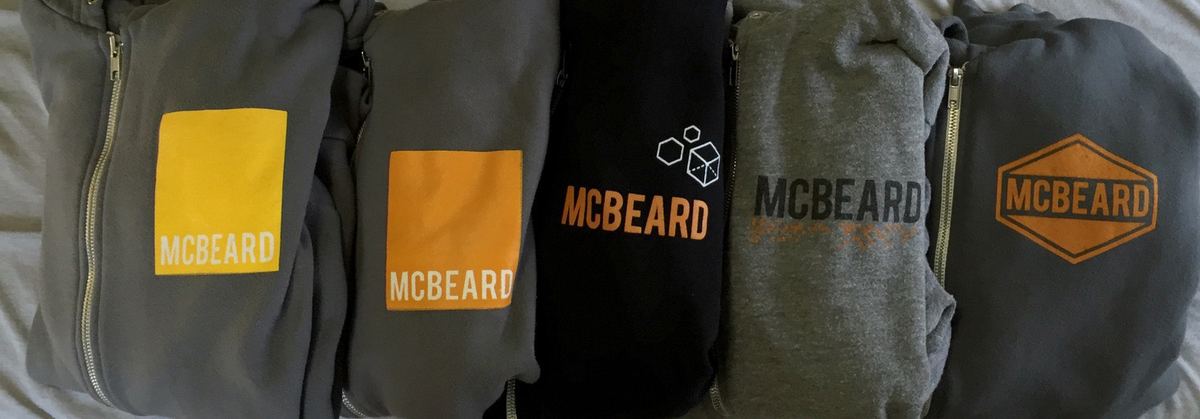 Five folded McBeard hoodies in a row: gray with a yellow block logo, gray with an orange block logo, black with an orange wordmark and cubes, heather gray with a distressed wordmark, and navy with an orange hexagon badge